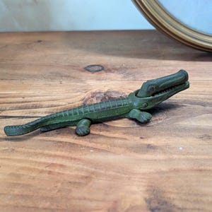 May include: A vintage green alligator-shaped nutcracker made of metal. The alligator's mouth is open, revealing the nut-cracking mechanism. The alligator is approximately 8 inches long.