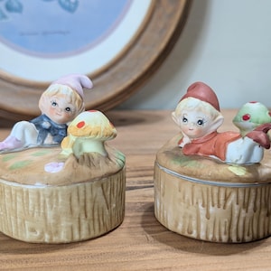 Könnte beinhalten: Zwei Keramik-Schmuckkästchen in Form von Baumstümpfen mit einer Fee oder Elfenfigur darauf. Eine Schachtel hat einen rot-behüteten Elf mit einem Pilz und die andere einen blau-behüteten Elf mit einem Pilz.