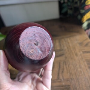 K&ouml;nnte beinhalten: Eine runde, dunkelrote Holzvase mit glatter, polierter Oberfl&auml;che. Der Boden der Vase zeigt die nat&uuml;rliche Holzmaserung und ein kleines Loch. Die Vase wird in der Hand einer Person gehalten.