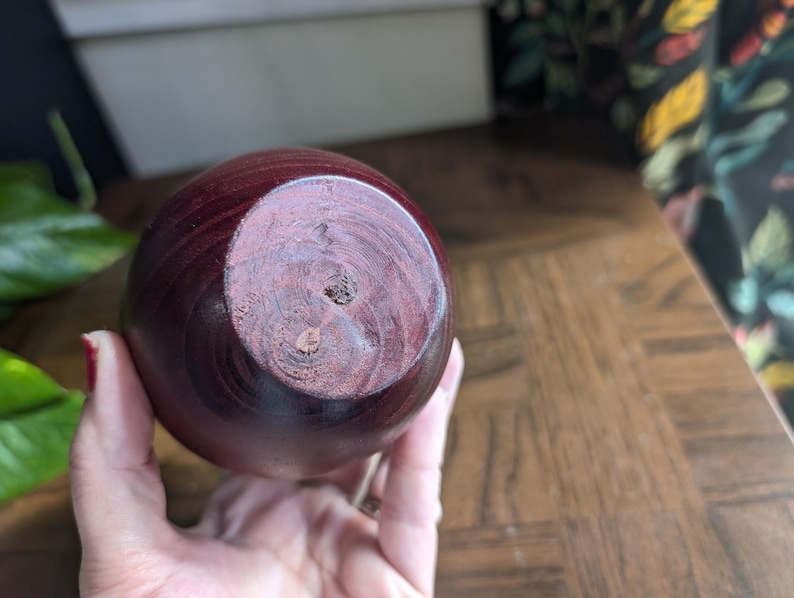 K&ouml;nnte beinhalten: Eine runde, dunkelrote Holzvase mit glatter, polierter Oberfl&auml;che. Der Boden der Vase zeigt die nat&uuml;rliche Holzmaserung und ein kleines Loch. Die Vase wird in der Hand einer Person gehalten.