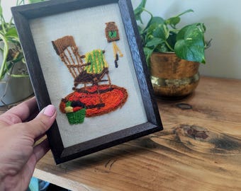 May include: A framed embroidery piece on off-white fabric featuring a brown rocking chair with a green blanket, a red circular rug, a small green basket with colorful items, and a hanging brown lantern with a green accent.