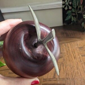 K&ouml;nnte beinhalten: Eine dekorative Apfelskulptur mit dunkelburgunderfarbenem Finish und einem silbernen Metallstiel und Bl&auml;ttern. Der Apfel ist rund und glatt, mit einem detaillierten Stieldesign. Die Skulptur steht auf einer Holzoberfl&auml;che.