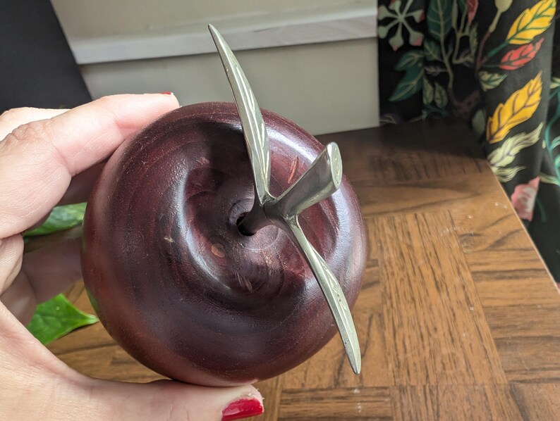 K&ouml;nnte beinhalten: Eine dekorative Apfelskulptur mit dunkelburgunderfarbenem Finish und einem silbernen Metallstiel und Bl&auml;ttern. Der Apfel ist rund und glatt, mit einem detaillierten Stieldesign. Die Skulptur steht auf einer Holzoberfl&auml;che.