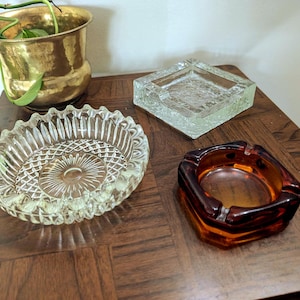 May include: Three vintage ashtrays on a wooden surface. One is clear glass with a diamond pattern, another is a textured square, and the third is a brown, square-shaped ashtray. A brass planter with greenery is in the background.