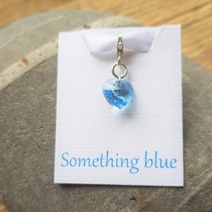 Puede incluir: Un pequeño charm de corazón de cristal azul en una cadena de plata. El charm está unido a una tarjeta blanca con el texto "something blue".