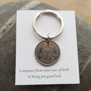 May include: A silver sixpence coin keychain with the year 1948 on it. The coin is attached to a silver key ring. A white card with the text "A sixpence from your year of birth to bring you good luck" is behind the coin.