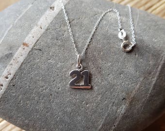 Puede incluir: Un collar de plata con un colgante con el número "21". El colgante cuelga de una delicada cadena. El collar se muestra sobre una superficie de piedra gris, con un fondo de madera.