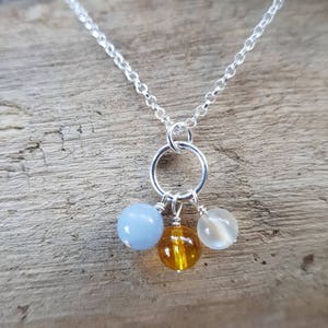 Peut inclure: Un collier en chaîne argent avec un pendentif rond en argent. Le pendentif a trois petites perles attachées : une bleu clair, une ambre et une blanche.