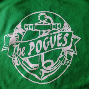 Op de afbeelding: Groen T-shirt met een witte afbeelding van een anker en de tekst "The Pogues".