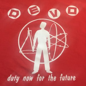 Op de afbeelding: Rood T-shirt met een wit silhouet van een persoon die voor een cirkel met geometrische vormen staat. De tekst "DEVO" staat bovenaan het shirt en de tekst "duty now for the future" staat onderaan.