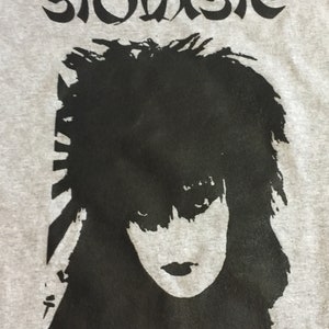 May include: A gray t-shirt with a black and white image of Siouxsie Sioux, a singer, with the word "Siouxsie" printed above the image.