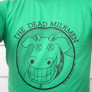 Könnte beinhalten: Grünes T-Shirt mit einem schwarz-weißen Grafikdruck einer lächelnden Kuh mit X-en als Augen. Die Grafik befindet sich in einem Kreis mit dem Text "The Dead Milkmen" darum herum.