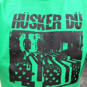 Pode incluir: Camiseta verde com o texto "Hüsker Dü" impresso em preto. A camiseta apresenta um gráfico em preto e branco de uma banda tocando em um palco com um fundo de bandeira americana.