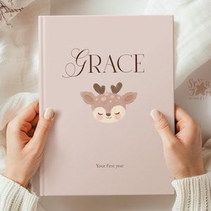 May include: A light pink baby book with the name "Grace" in elegant script. A cartoon deer with heart-shaped antlers and closed eyes is below the name. The text "Your first year" is at the bottom.