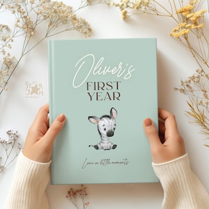 May include: A light teal baby memory book with the text "Oliver's FIRST YEAR" in white and brown script. A watercolor illustration of a zebra cub is centered below the text. The phrase "Love in little moments" is written at the bottom.
