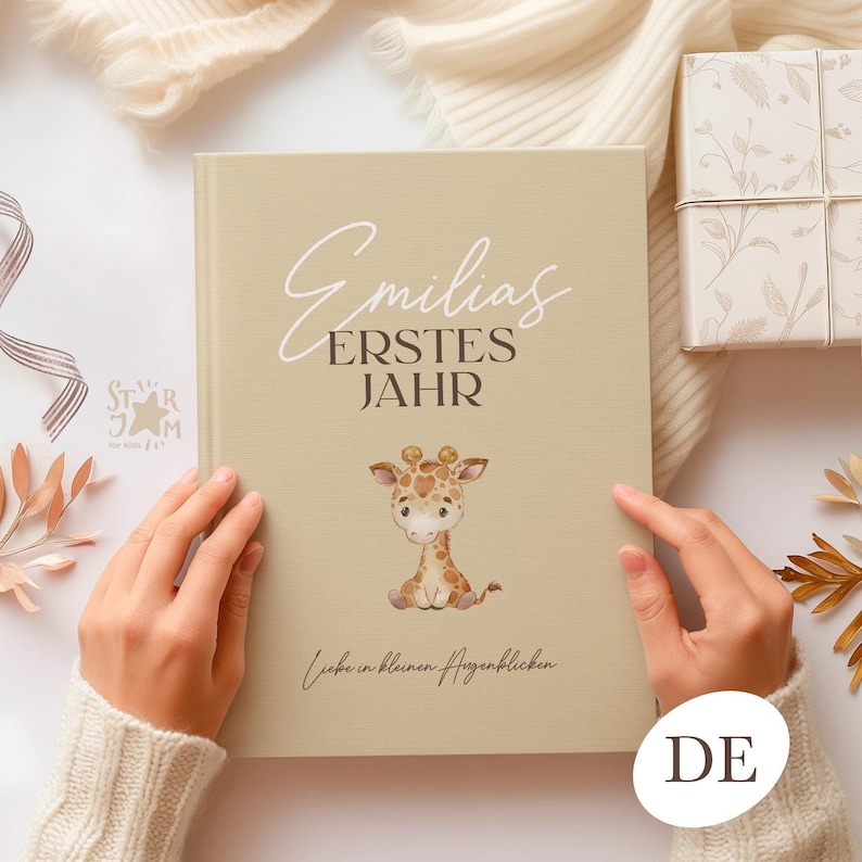 以下が含まれることがあります： ベージュ色のベビーメモリーブック。テキスト「Emilias ERSTES JAHR」とキリンのイラストが描かれています。本は両手で開かれています。本の底には「Liebe in kleinen Augenblicken」と書かれています。