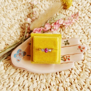May include: A gold ring with a pink oval gemstone and two smaller white gemstones on either side, displayed in a yellow velvet ring box.