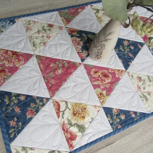 Könnte beinhalten: Gesteppter Tischläufer mit floralem Muster in Rot, Creme und Marineblau. Der Läufer besteht aus dreieckigen Stoffstücken mit weißen Steppakzenten. Die Kanten sind mit einem blau gestreiften Rand versehen. Eine dekorative Vase befindet sich darauf.