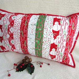 May include: A rectangular Christmas-themed pillow with a red border. The pillow features a patchwork design with alternating patterns of snowmen, candy canes, and polka dots in red, white, and green. The pillow is resting on a white surface.