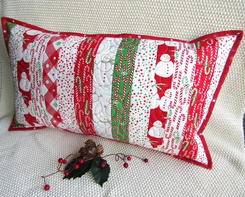 May include: A rectangular Christmas-themed pillow with a red border. The pillow features a patchwork design with alternating patterns of snowmen, candy canes, and polka dots in red, white, and green. The pillow is resting on a white surface.