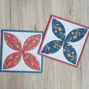 Könnte beinhalten: Zwei quadratische, gesteppte Tischsets. Eines hat einen blauen Rand und rote Blütenblätter. Das andere hat einen roten Rand und blaue Blütenblätter. Beide haben weiße Zentren mit floralen Mustern.