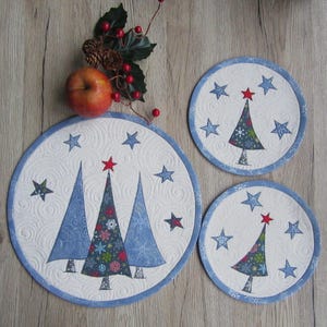Può includere: Set di tre tovagliette natalizie trapuntate a forma di albero di Natale. Ogni tovaglietta presenta uno sfondo bianco con stelle blu e un bordo blu. La tovaglietta più grande ha tre alberi di Natale blu.