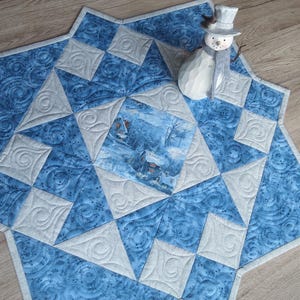 Könnte beinhalten: Ein sternförmiger, gesteppter Tischläufer mit einer Winterszene in der Mitte. Der Läufer hat ein blau-weißes Farbschema mit einer Schneemannfigur oben drauf. Der Quilt hat ein detailliertes Muster.