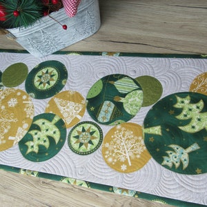 Pode incluir: Um caminho de mesa de Natal acolchoado com desenhos circulares verdes e dourados com padrões de árvores e flocos de neve. O caminho de mesa tem um fundo branco com um padrão de redemoinho e uma borda verde. Um balde decorativo com vegetação está no fundo.