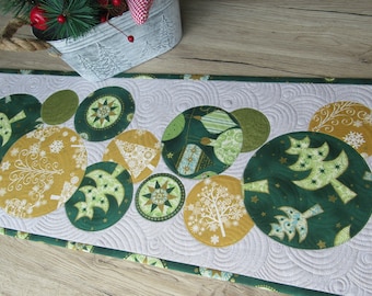 May include: A quilted Christmas table runner featuring green and gold circular designs with tree and snowflake patterns. The runner has a white background with a swirl pattern and a green border. A decorative bucket with greenery is in the background.