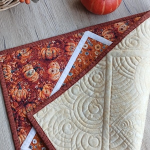 May include: A quilted table runner featuring a vibrant orange and brown pumpkin print on one side and a solid cream-coloured fabric with intricate, swirling machine-stitched patterns on the reverse side, finished with a dark brown fabric border.