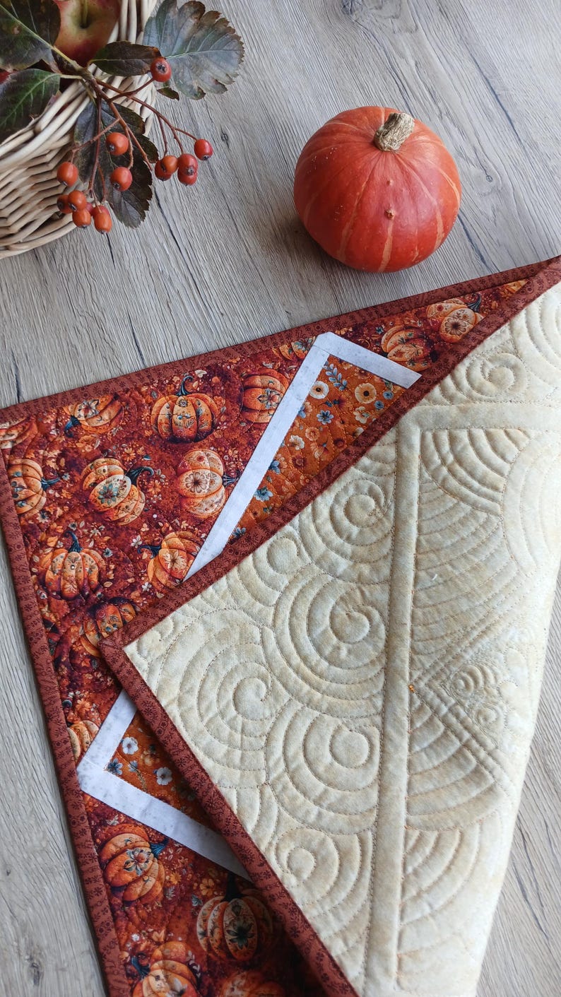May include: A quilted table runner featuring a vibrant orange and brown pumpkin print on one side and a solid cream-coloured fabric with intricate, swirling machine-stitched patterns on the reverse side, finished with a dark brown fabric border.