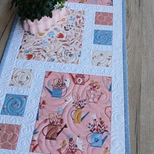 Könnte beinhalten: Quilt-Tischläufer mit Blumen- und Gießkannenmuster in Rosa, Blau und Weiß. Der Läufer hat einen hellblauen Rand mit weißen Punkten und ein dekoratives Blumenarrangement.