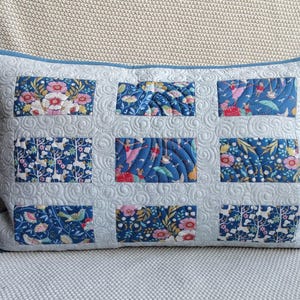 May include: Rectangular decorative pillow with a light blue quilted background and a dark blue border. The pillow features nine rectangular patches with floral and whimsical designs in shades of blue, pink, and white. The pillow is approximately 20 inches long.