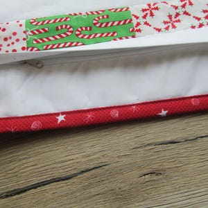 May include: Close-up of a white pillowcase with a zipper, featuring a patchwork design with red polka dots, candy canes, and snowflakes. The edge is trimmed with a red fabric with white stars and swirls. The pillowcase is on a wooden surface.