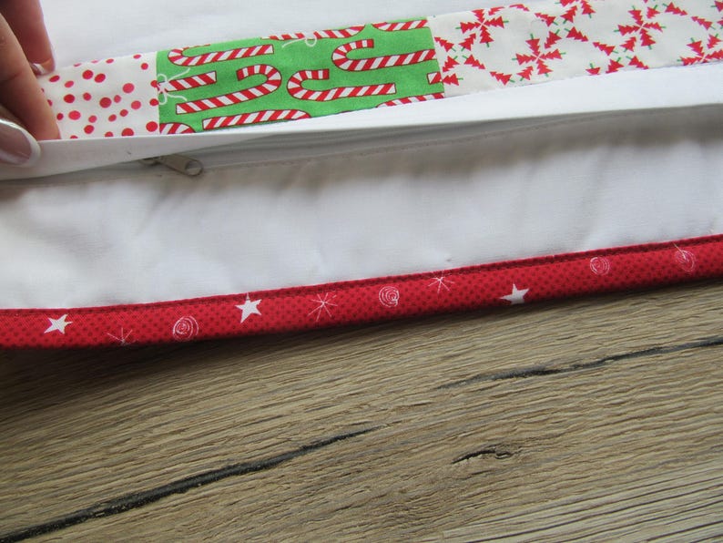 May include: Close-up of a white pillowcase with a zipper, featuring a patchwork design with red polka dots, candy canes, and snowflakes. The edge is trimmed with a red fabric with white stars and swirls. The pillowcase is on a wooden surface.