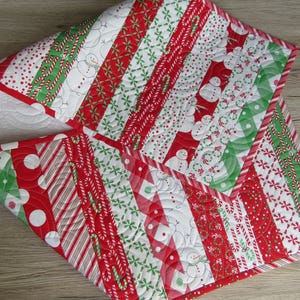 May include: A Christmas-themed quilted table runner with a patchwork design. The runner features alternating stripes of red, white, and green fabrics with patterns including snowmen, candy canes, and polka dots. The edges are finished with a red border.