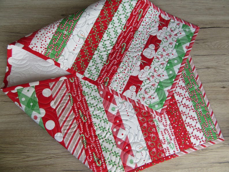 May include: A Christmas-themed quilted table runner with a patchwork design. The runner features alternating stripes of red, white, and green fabrics with patterns including snowmen, candy canes, and polka dots. The edges are finished with a red border.