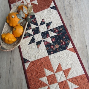 May include: A quilted table runner with star patterns in shades of rust, cream, and navy blue. The runner features intricate stitching and a floral fabric accent. A basket of small orange pumpkins and a decorative gourd are placed on the wooden surface.