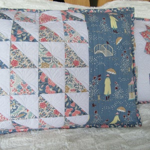 May include: Two decorative pillows with floral patterns. One pillow has a blue background with a white bridge and people walking in the rain. The other pillow has a white background with a patchwork of floral fabric triangles.
