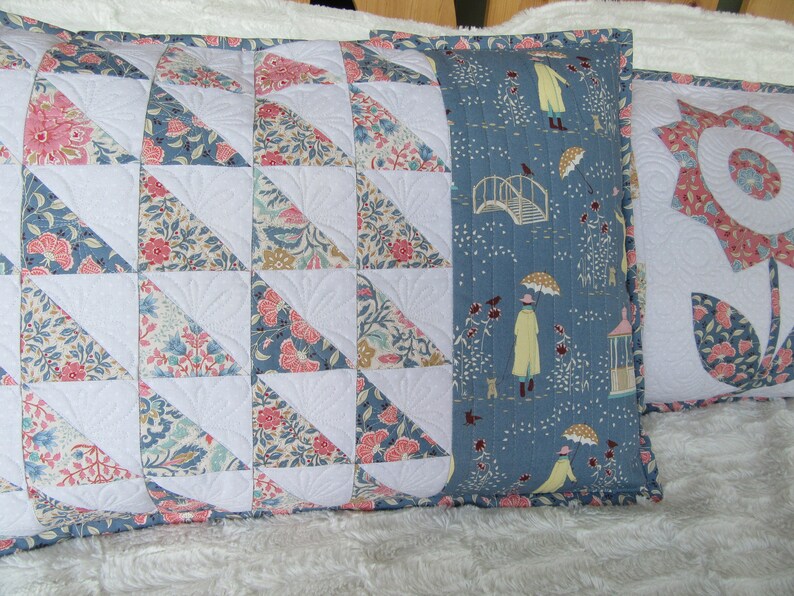 May include: Two decorative pillows with floral patterns. One pillow has a blue background with a white bridge and people walking in the rain. The other pillow has a white background with a patchwork of floral fabric triangles.