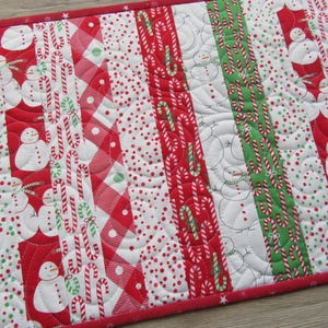 May include: A quilted Christmas table runner with a red border. The design features alternating vertical stripes of snowmen, candy canes, and polka dots in red, white, and green. The runner is approximately 12 inches wide.
