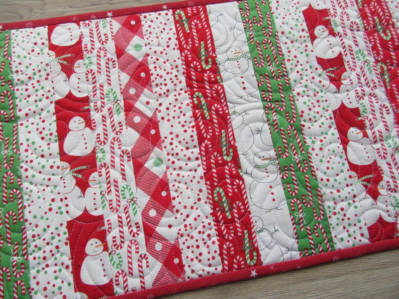 May include: A quilted Christmas table runner with a red border. The design features alternating vertical stripes of snowmen, candy canes, and polka dots in red, white, and green. The runner is approximately 12 inches wide.