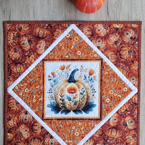 May include: A square quilted fabric mat featuring a central white panel with a decorative pumpkin illustration surrounded by floral motifs. The mat has a diamond-shaped border and an outer field patterned with small pumpkins and flowers in orange tones.