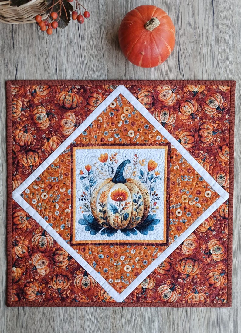 May include: A square quilted fabric mat featuring a central white panel with a decorative pumpkin illustration surrounded by floral motifs. The mat has a diamond-shaped border and an outer field patterned with small pumpkins and flowers in orange tones.