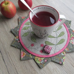 K&ouml;nnte beinhalten: Ein grauer und rosa gesteppter Untersetzer mit Blumenmuster. Eine wei&szlig;e Tasse mit dem Text "Gl&uuml;ckmomente" steht auf dem Untersetzer. Die Tasse ist mit einer roten Fl&uuml;ssigkeit gef&uuml;llt und ein silberner L&ouml;ffel befindet sich darin.