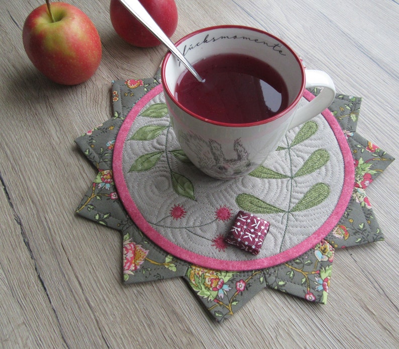 K&ouml;nnte beinhalten: Ein grauer und rosa gesteppter Untersetzer mit Blumenmuster. Eine wei&szlig;e Tasse mit dem Text "Gl&uuml;ckmomente" steht auf dem Untersetzer. Die Tasse ist mit einer roten Fl&uuml;ssigkeit gef&uuml;llt und ein silberner L&ouml;ffel befindet sich darin.