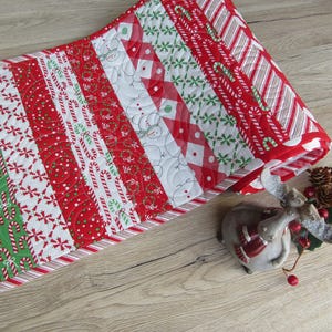 Könnte beinhalten: Ein weihnachtlicher Tischläufer mit Patchwork-Design. Der Läufer hat rote, weiße und grüne Stoffe mit Zuckerstangen-, Schneeflocken- und Schneemannmustern. Der Läufer ist etwa 30 cm breit und 91 cm lang.