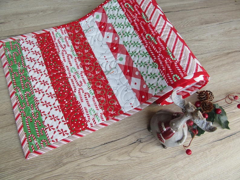 May include: A Christmas-themed table runner with a patchwork design. The runner features red, white, and green fabrics with candy cane, snowflake, and snowman patterns. The runner is approximately 12 inches wide and 36 inches long.