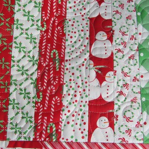 May include: A festive Christmas quilt with alternating vertical stripes. The quilt features red and white candy canes, snowmen, snowflakes, and wreaths. The quilt is bordered with a red and white striped pattern.