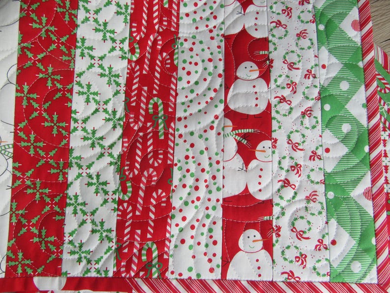 May include: A festive Christmas quilt with alternating vertical stripes. The quilt features red and white candy canes, snowmen, snowflakes, and wreaths. The quilt is bordered with a red and white striped pattern.